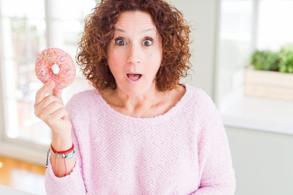 Senior woman eating pink sugar donut scared in shock with a surprise face, afraid and excited with fear expression